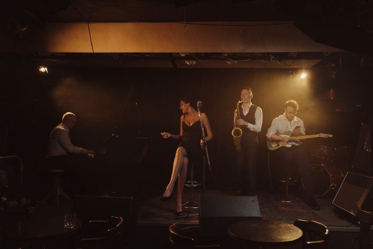 A singer, pianist, saxophonist, and guitarist performing on a small stage under a warm spotlight